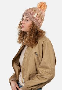 Knit beanie in pink and orange hues with a pom-pom on top; features textured patterns. Worn with a tan bomber jacket and cream sweater.