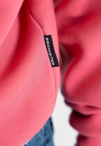 Gros plan d’un vêtement rose avec une étiquette en tissu noire portant la mention "PROHIBITED", cousue dans la couture latérale près de la taille.