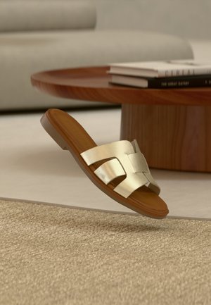 Singolo sandalo a ciabatta color cuoio e oro con cinturini traforati che fluttua sopra un tappeto beige strutturato accanto a un tavolo rotondo in legno.