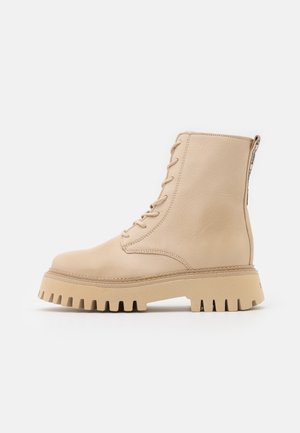 Bottine beige à lacets avec semelle épaisse à crampons, texture en cuir lisse et languette à l’arrière sur fond blanc.