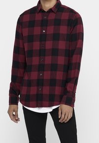 Punamusta ruudullinen flanellipaita, jossa pitkät hihat, napillinen edessä ja vino helma, kerroksittain valkoisen paidan päällä. Kankaan rakenne on teksturoitu.