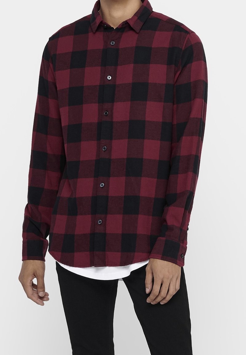 Punamusta ruudullinen flanellipaita, jossa pitkät hihat, napillinen edessä ja vino helma, kerroksittain valkoisen paidan päällä. Kankaan rakenne on teksturoitu.