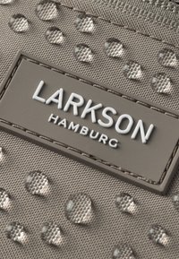 Szürke textúrájú szövet, kiemelkedő vízcseppekkel, egy téglalap alakú címkével, amelyen fehér betűkkel a "LARKSON HAMBURG" felirat szerepel.