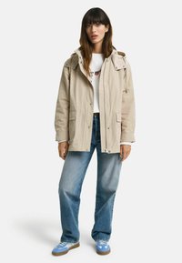 Beige hættetrøje med trykknaplukninger og to forlommer, parret med lyseblå jeans og blå sneakers med grå detaljer.