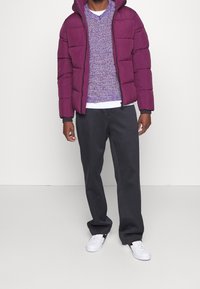 Puchowa fioletowa kurtka z kapturem, nałożona na fioletowy sweter o fakturze, zestawiona z ciemnymi spodniami i białymi trampkami. Prosty i nowoczesny design.