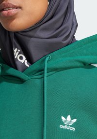 Grön hoodie med en texturerad tyg, med en vit Adidas-logotyp på bröstet och en mörkgrå mönstrad hijab under huvan.