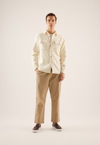 Cream-colored button-up shirt with two chest pockets, paired with light brown trousers and brown slip-on shoes. Plain background.