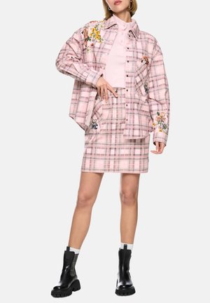 Woman wearing a pink plaid skirt and matching oversized shirt with floral embroidery, pale pink top, white socks, and black boots.
