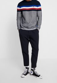 Marineblaues Strickpullover mit blauen, roten und weißen Streifen, kombiniert mit maßgeschneiderten dunkelgrauen Hosen und schwarzen High-Top-Sneakers mit weißen Sohlen.