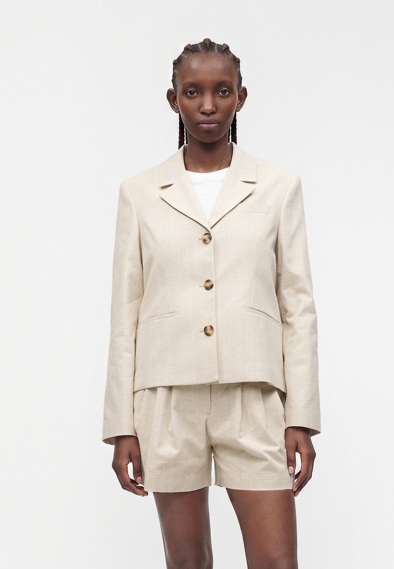 Femme aux cheveux tressés portant un blazer beige à trois boutons sur un top blanc et un short plissé taille haute assorti.