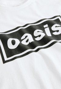 Tissu blanc avec un logo rectangulaire noir comportant le mot "oasis" en lettres minuscules blanches à l’intérieur du rectangle.