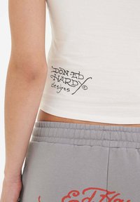 Bílý přiléhavý crop top s černým textem "Don Ed Hardy designs" na boku, v kombinaci s šedými tepláky s elastickým pasem a červeným grafickým potiskem.