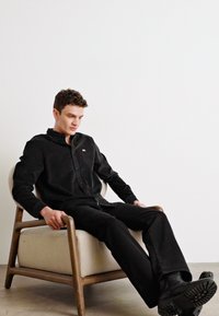 Camisa de pana negra y pantalones a juego que lleva puesta una persona sentada, con una silla de madera clara y un fondo blanco liso.