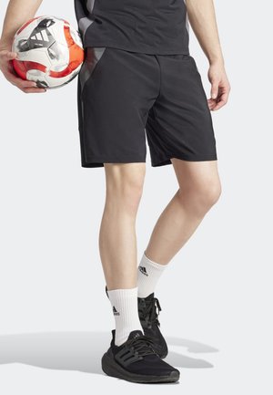 Pantaloni sportivi neri con pannelli laterali, abbinati a calze bianche a metà polpaccio e sneakers nere. Un pallone da calcio rosso e bianco è tenuto nella mano sinistra.