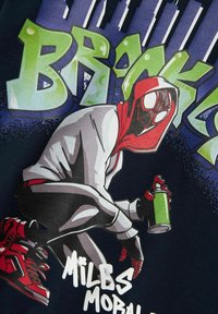 Zwarte shirt met een gehurkte figuur in een rode hoodie met een spinnenwebontwerp, die een groene spuitbus vasthoudt, met graffiti-achtige tekst erachter.