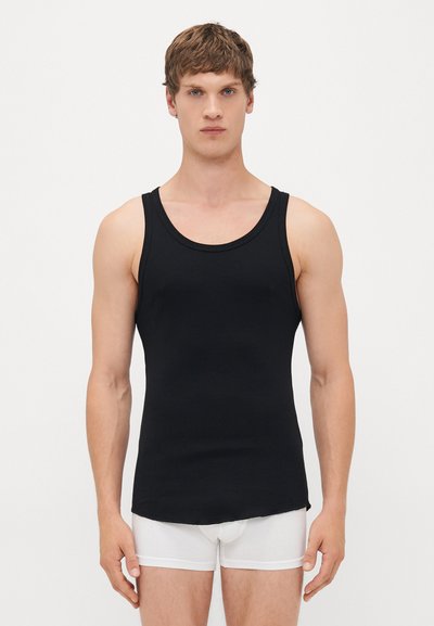 Mann in einem enganliegenden schwarzen Tanktop und weißen Boxershorts, der vor einem schlichten weißen Hintergrund steht und nach vorne blickt.