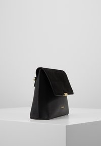 Černá kožená kabelka s texturovaným povrchem a semišovým klopou. Obsahuje zlatý zapínací mechanizmus. Úhlový pohled ukazuje její trojúhelníkový tvar.