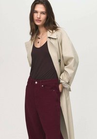 Béžový trench coat přes hnědé tílko, doplněný vínovými kalhotami s vysokým pasem. Mezi výrazné detaily patří zapínání na knoflíky a vrstvený náhrdelník.