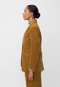 Mosterdkleurige ribfluwelen blazer met gestructureerde revers, zijzakken en knoopmanchetten, gecombineerd met een grijze coltrui. Zijprofielweergave.