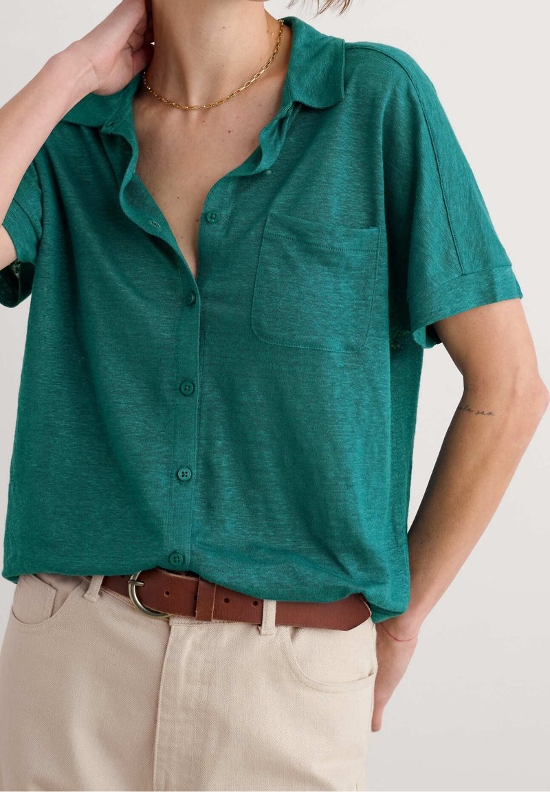 Personne portant une chemise bleu sarcelle à manches courtes et à boutons, un pantalon beige avec une ceinture marron, un collier en chaîne dorée, main gauche touchant le cou.
