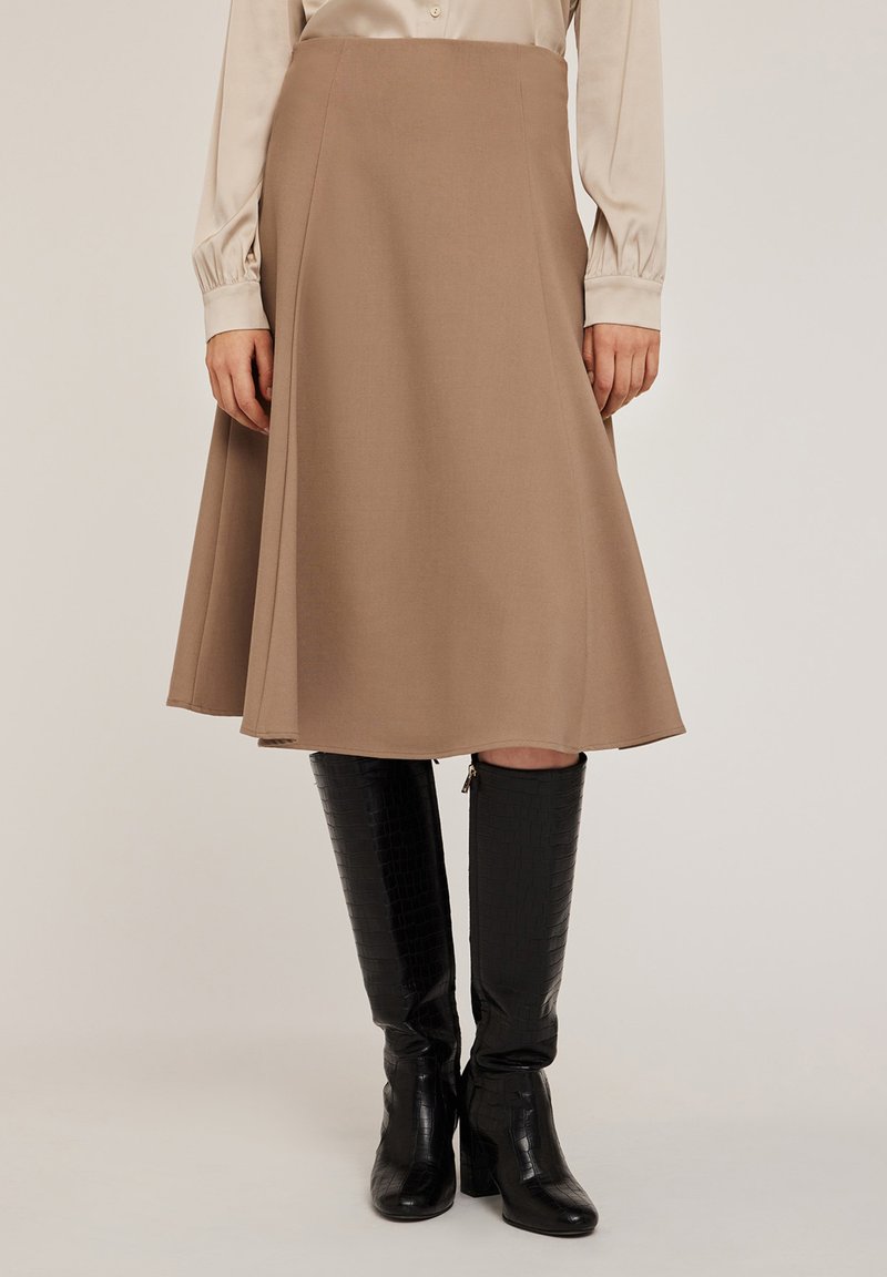 Una gonna a trapezio di lunghezza al ginocchio in tessuto marrone chiaro con una trama liscia, abbinata a stivali neri alti, lucidi, in pelle stampata coccodrillo.