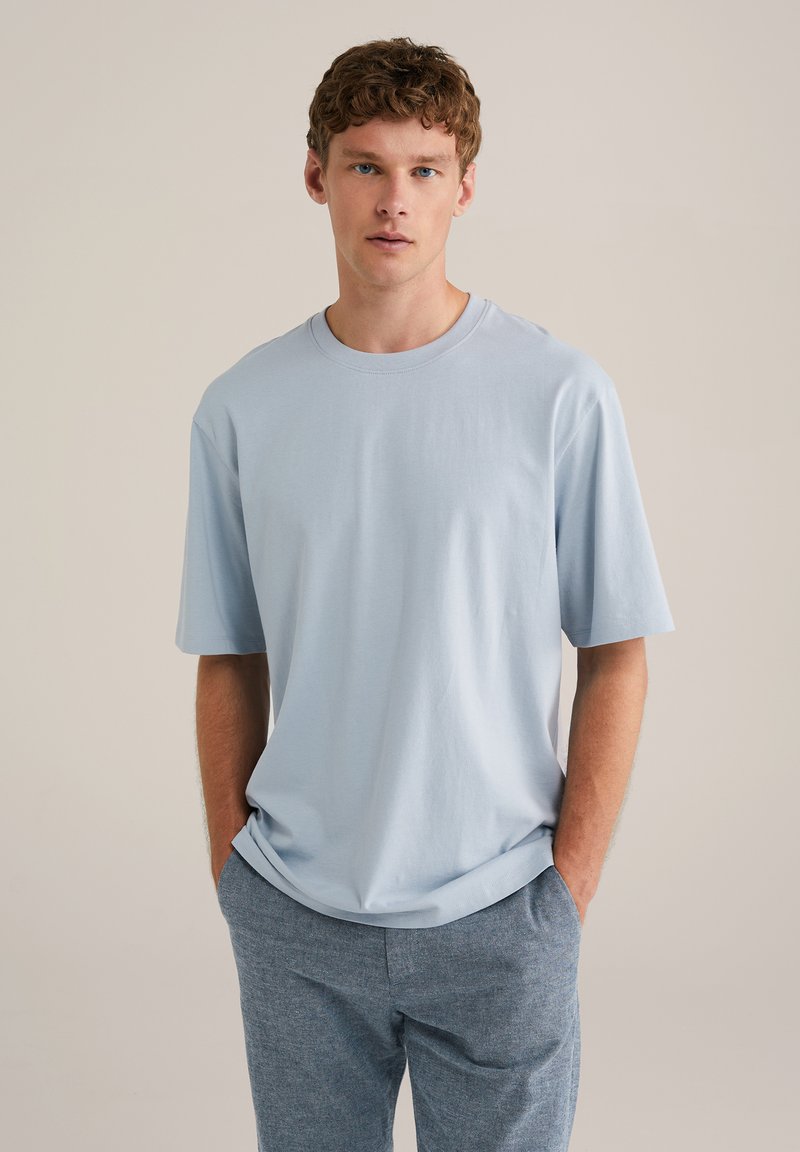Himmelblaues Baumwoll-T-Shirt mit kurzen Ärmeln, Rundhalsausschnitt, lockere Passform, glatte Textur. Kombiniert mit grauen gemusterten Hosen.