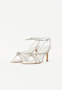 Silver strappy high-heeled sandals with a knot detail at the front, featuring transparent straps and a slender heel. Soft suede texture.
