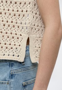 Gros plan sur un bras portant un haut en crochet crème avec une fente sur le côté, sur un jean en denim bleu clair.