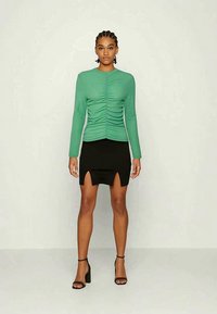 Mulher com cabelo trançado vestindo uma blusa verde de mangas longas com drapeado, uma saia mini preta com aberturas frontais e sandálias de salto alto pretas, em pé.