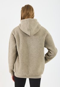 Bež flis dukserica s teksturiranom izradom, oversized krojem i kenguruh džepom. Ima kapuljaču s užetom i rebraste manžete.