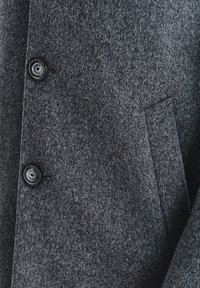 Szary wełniany płaszcz o fakturowanej tkaninie, z dwoma czarnymi guzikami i detalem bocznej kieszeni. Projekt jest dopasowany z czystym wykończeniem.