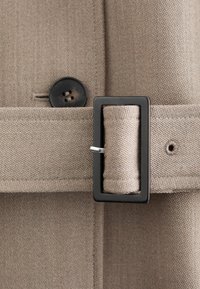 Beige stofbælte med sort rektangulært spænde fastgjort omkring en matchende beige tekstureret frakke med en synlig sort knap.