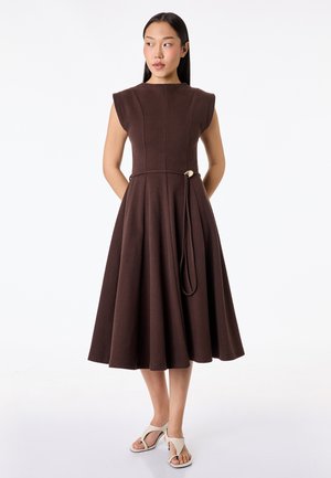 Femme portant une robe midi marron foncé sans manches avec des épaules structurées et des talons blancs ouverts, debout devant un fond blanc.