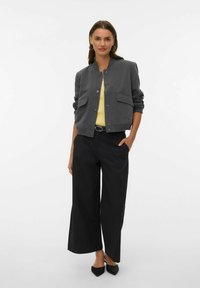 Szara kurtka bomberka z zapięciem na guziki i dwoma przednimi kieszeniami, zestawiona z żółtą bluzką i czarnymi szerokimi spodniami, na białym tle.