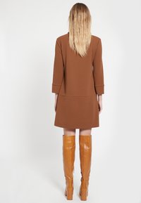 Brun skräddarsydd klänning med trekvartslånga ärmar, ovanför knäet, bärs med knähöga tan läderstövlar. Slät textur, minimal detaljering.