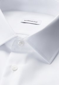 Weißes gefaltetes Hemd mit spitzem Kragen und zwei weißen Knöpfen, mit einem sichtbaren Markenetikett, das innen im Kragen eingenäht ist.