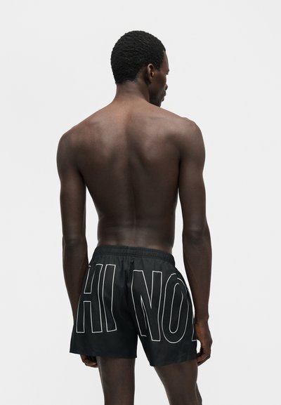 Άνδρας από πίσω που φοράει μαύρα σορτς με μεγάλα λευκά γράμματα με περίγραμμα στην πλάτη, στέκεται μπροστά σε απλό φωτεινό φόντο.