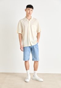 Camicia giallo chiaro a maniche corte, con texture e tasca, abbinata a pantaloni corti in denim blu chiaro e sneakers bianche.