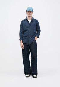 Hombre joven con camisa de botones a rayas azules, jeans oscuros anchos, mocasines blancos y negros, y una gorra azul clara, de pie con una mano en el bolsillo.