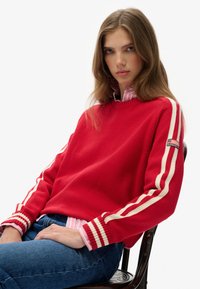 Jonge vrouw met lang bruin haar die een rode trui met witte strepen, een roze gestreept shirt en een blauwe spijkerbroek draagt, terwijl ze op een stoel zit.