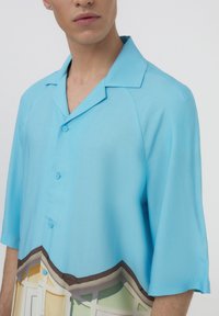 Camicia a maniche corte in tessuto blu chiaro con colletto a bottoni. Presenta una stampa colorata di case lungo l'orlo e una texture liscia.