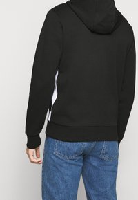 Felpa nera con cappuccio e tasca a marsupio, presenta inserti bianchi laterali e cappuccio con coulisse, realizzata in tessuto morbido, abbinata a jeans blu.