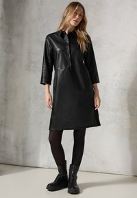 Schwarzes Lederkleid mit Kragen, Dreiviertelärmel und Knopfleiste. Glatte Textur, leicht A-Linie Form, getragen mit schwarzen Knöchelstiefeln.