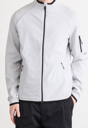 Uomo che indossa una giacca a zip integrale grigio chiaro con rifiniture nere e tasche con cerniera, abbinata a pantaloni neri, in piedi con le mani rilassate.