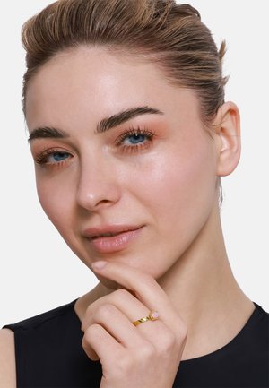 Goldring mit verdrehter Band und quadratischem Stein. Die Haut hat ein glattes, natürliches Finish; die Haare sind nach hinten gestylt. Schwarzes Oberteil mit rundem Ausschnitt.