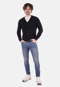Cárdigan navy de cuello en V, mangas largas y cierre de botones. Llevado sobre una camisa blanca, combinado con jeans azules y zapatillas.