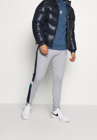 Giacca imbottita blu navy, felpa blu con cappuccio, pantaloni pantaloni grigi con strisce laterali blu navy e teal, e sneakers bianche con accenti neri.