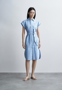 Ženska stoji oblečena v svetlo modro obleko brez rokavov z gumbi, s pasom, povezanem v pasu, in belimi sandali na sivi podlagi ob preprosti svetli steni.