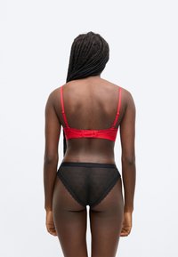 Piros szatén melltartó állítható pántokkal, csipkével díszítve; fekete, áttetsző alsóneművel párosítva, amelyen ismétlődő logóminta és hullámos szegély található.