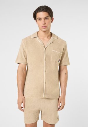 Ung mand iført beige kortærmet skjorte med knapper og matchende shorts med lille logo på lommen, stående mod en ensfarvet baggrund.